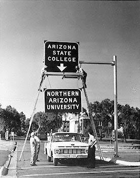 A black and white photograph captured in 1966 of the new interstate sign reading "Northern Arizona University" being installed to replace the previous sign reading "Arizona State Colleg"