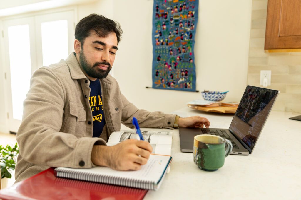 N A U student studying at home, taking notes while working on a laptop.