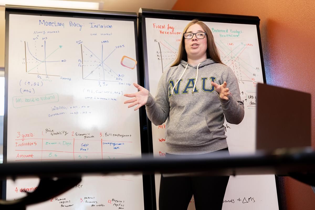 An economics student stands in front of a whiteboard with charts and graphs.