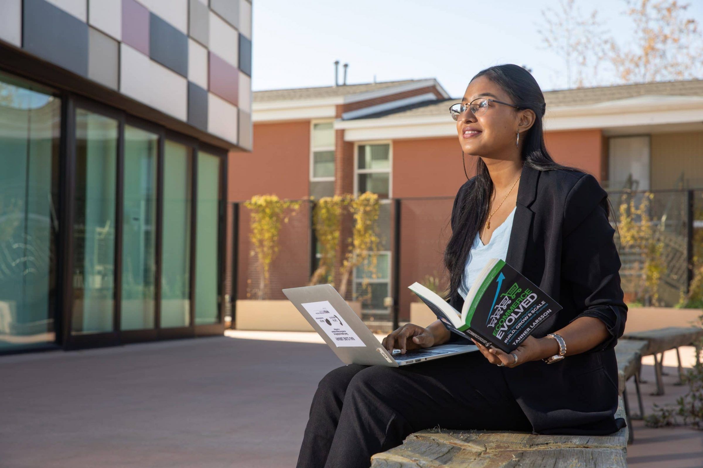 Student outside working on a laptop while holding a book.